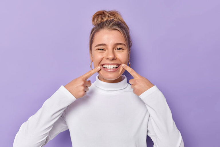 Happy woman in white sweater pointing at her teeth and smiling against a purple background, showcasing a healthy and confident smile.